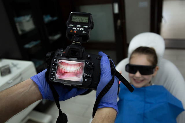 Camera in gloved hands showing a retracted smile photo on its screen, patient in the dental chair behind